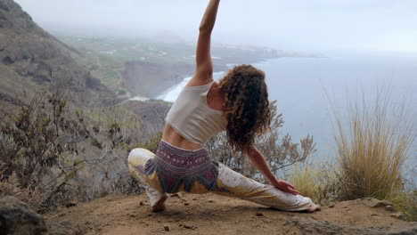 Perched-on-a-cliff's-edge,-a-woman-assumes-a-warrior-pose,-raising-her-arms-and-breathing-in-the-ocean-air-as-she-practices-yoga